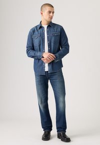 Camicia in denim blu scuro con due tasche sul petto, bottoni a pressione e cuciture a contrasto, abbinata a jeans blu più chiari e stivali neri.