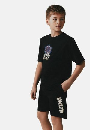 Chlapec nosiaci čierne tričko s fialovou smajlíkovou tvárou a textom, a čierne šortky s bielym nápisom, stojaci s jednou rukou v vrecku.