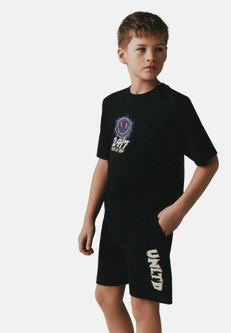 Chlapec v černém tričku s fialovým smajlíkem a nápisem a v černých šortkách s bílým nápisem, stojící s jednou rukou v kapse.