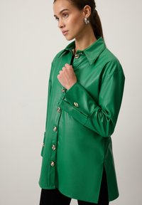 Zaļš mākslīgās ādas krekls ar norādītu apkakli, zelta pogu aizdari un sānu spraugām. Gluda tekstūra, garas piedurknes un brīva piegriezuma.