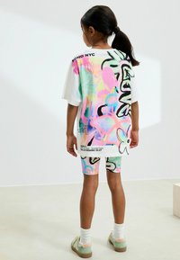 T-shirt taille oversize blanche avec des motifs de graffiti colorés et du texte, assortie avec un short. Comprend des tons pastel et des accents graphiques.