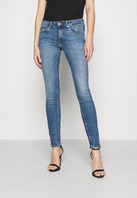 Jeans ajustados azules fabricados en denim, con cinco bolsillos, cierre de botón metálico y un ligero desgaste. Llevados con tacones negros con tiras.