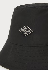 Zwarte bucket hat van gladde stof, voorzien van een opvallende zilveren diamanten logo patch met de tekst "REPLAY" en een geribbelde randtextuur.