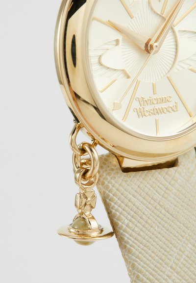 Χρυσό ρολόι με ανάγλυφη κρέμα ζώνη, που διαθέτει καντράν με ηλιακό σχέδιο και ένα κρεμαστό γούρι σε σχήμα πλανήτη με μια χάντρα.