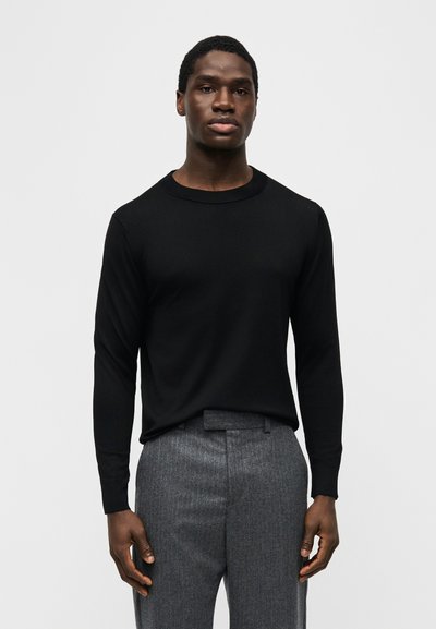 Homme debout, faisant face à l'avant, portant une chemise noire à manches longues et un pantalon gris cintré, sur un fond clair uni.