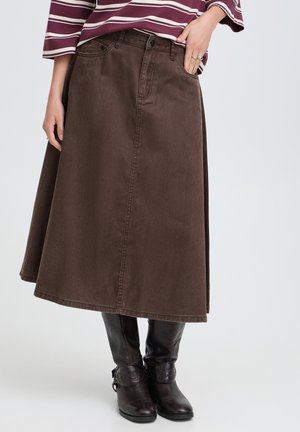 Femme portant une jupe midi marron en ligne A, un haut rayé et des bottes noires montant jusqu'aux genoux, debout avec une main dans la poche de la jupe.