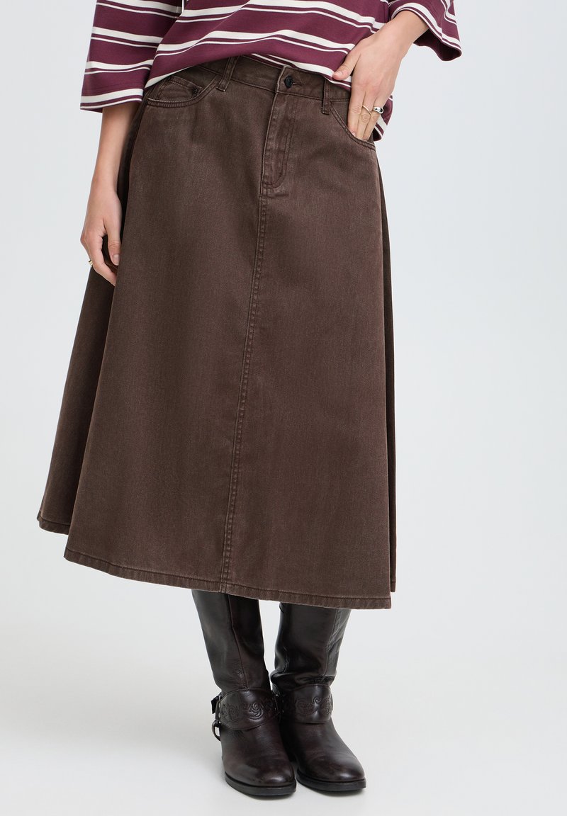 Femme portant une jupe midi marron en ligne A, un haut rayé et des bottes noires montant jusqu'aux genoux, debout avec une main dans la poche de la jupe.