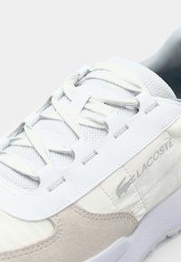 Lacoste sneaker in white and beige. Features mesh upper, flat laces, and rubber outsole. Logo on the side, textured accents on material.