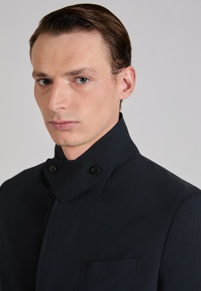 Man met naar achteren gekamd haar, gekleed in een zwarte jas met een hoge kraag en twee zichtbare knopen op de kraag, tegen een effen achtergrond.