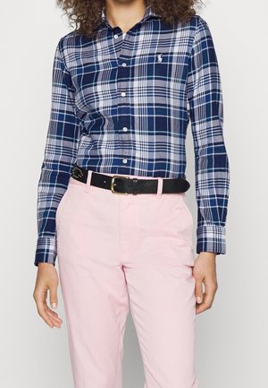 Person trägt ein Marineblau-weiß kariertes Hemd mit Knöpfen, das in eine hellrosa Hose gesteckt ist, und einen schwarzen Gürtel mit goldener Schnalle.