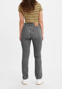 Jeans grises de cintura alta con pierna ajustada, confeccionados en tela de mezclilla. La vista trasera muestra dos bolsillos traseros y un parche de cuero con el logo en la cintura.