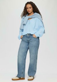 Lichtblauwe blouse met een ronde hals, gecombineerd met lichtblauwe jeans met een brede pijp. Een grijze trui over de schouders gedrapeerd. Beige en zwarte schoenen.