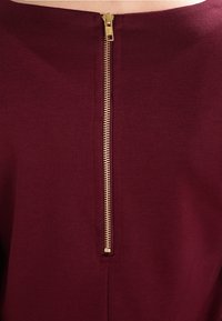 Maroon tyg topp med en bakre gyllene dragkedja med slät yta, kompletterad med en ren halsringning och strömlinjeformat design.