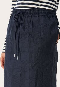 Donkerblauwe, gekreukelde rok met een elastische tailleband en trekkoord. Beschikt over zijzakken en een gestructureerde afwerking.