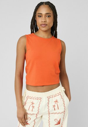 Femme aux cheveux tressés portant un crop top orange sans manches et un pantalon blanc avec une broderie de palmiers orange, mains dans les poches.