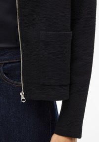 Μαύρο μάλλινο μπουφάν με φερμουάρ στο μπροστινό μέρος, πλαϊνή τσέπη και εφαρμοστή γραμμή. Το υλικό φαίνεται μαλακό με ριμπ μοτίβο.