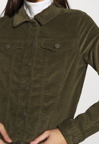Olive green corduroy jacket featuring a button-up front, collar, and two chest pockets with metal snap buttons. Vertical ribbed texture.