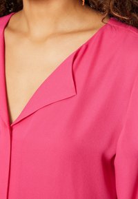 Blouse rose avec un décolleté en V, texture douce et légère drapée. Présente un design épuré sans embellissements ni motifs.