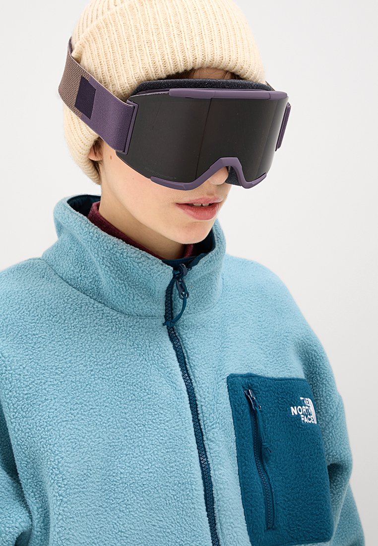 Brille mit dunklen Gläsern und lila Rahmen, getragen mit einer hellblauen Fleecejacke, die eine dunkelblaue Tasche hat. Strickmütze in Beige auf dem Kopf.