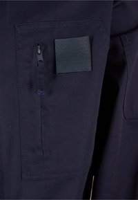 Tissu bleu marine foncé avec une poche zippée verticale et un patch carré en velcro cousu à côté, mettant en valeur les caractéristiques de rangement sur les vêtements.