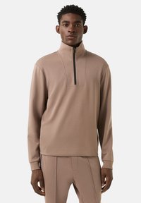 Béžový pulovr se čtvrtým zipem vpředu, dlouhými rukávy a hladkou texturou. Má jemný designový prvek na límci a přiléhavé manžety.