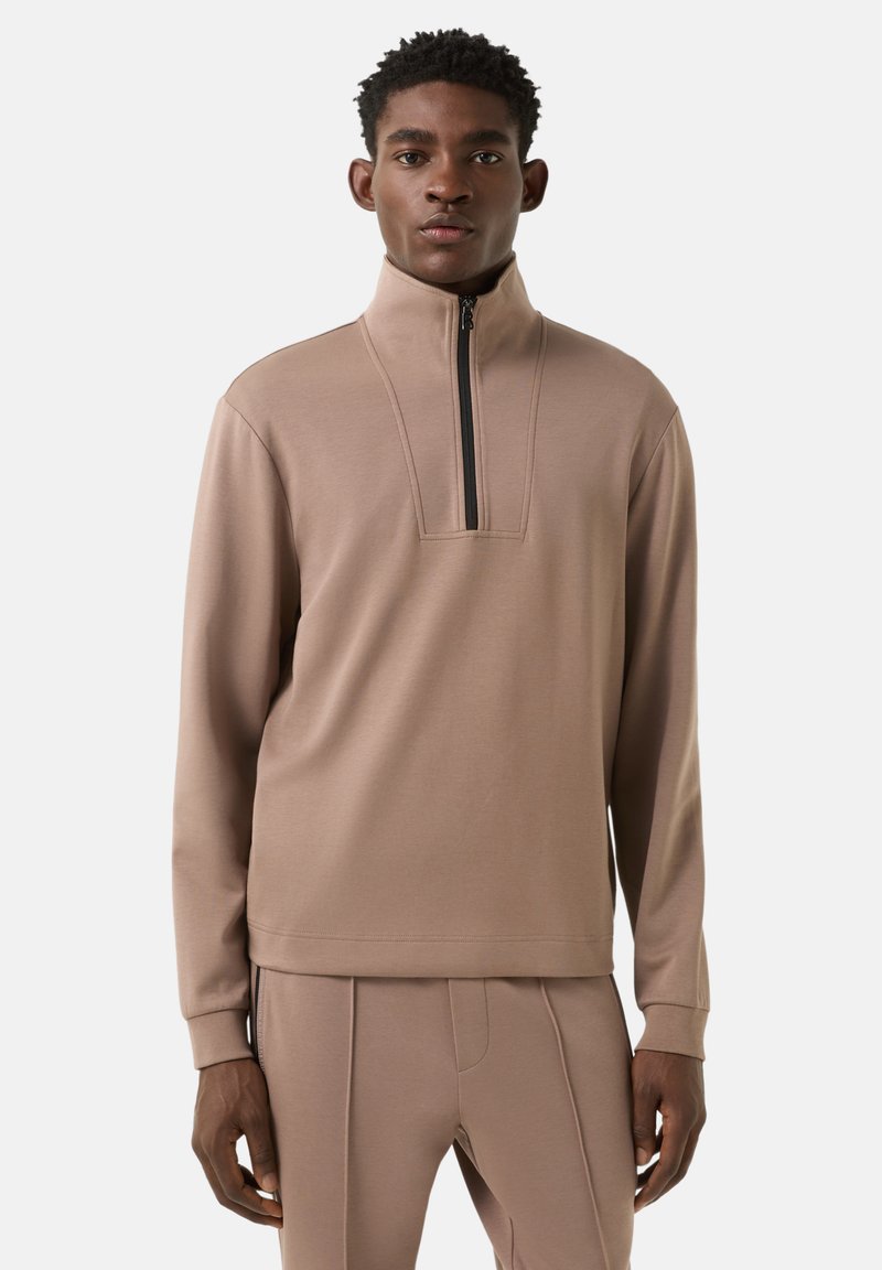 Béžový pulovr se čtvrtým zipem vpředu, dlouhými rukávy a hladkou texturou. Má jemný designový prvek na límci a přiléhavé manžety.