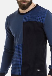 Blauer Pullover mit verschiedenen Texturen: gerippte Einsätze, Hahnentritt-Muster und einem einfarbigen, dunkelblauen Abschnitt. Rundhalsausschnitt, taillierte Passform.
