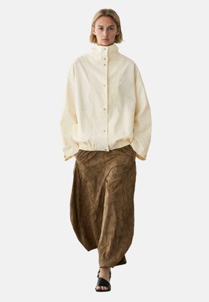 Frau mit kurzen blonden Haaren, die eine cremefarbene Jacke mit hohem Kragen, lockere braune strukturierte Hose und schwarze offene Sandalen trägt, geht vorwärts.