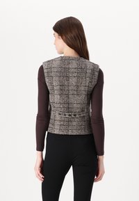 Brūns svītrains īsais vestes pieces no auduma tweed ar noapaļotu kakla izgriezumu un pogu detaļu aizmugurē, saskaņots ar garām piedurknēm tumšu augšu.