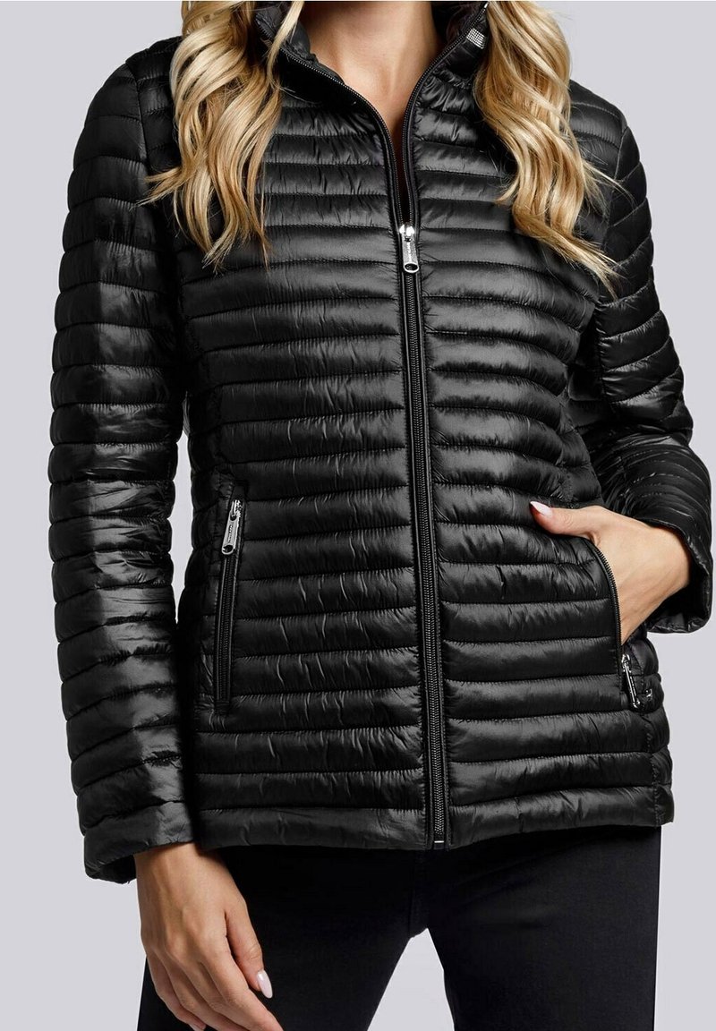 Femme portant une veste matelassée noire zippée avec des poches latérales zippées et une main dans une poche.