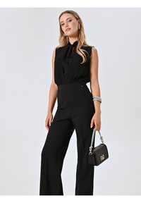 Zwart jumpsuit met mouwloos ontwerp, strikdetail bij de hals en wijde pijpen. Gecombineerd met een kleine zwarte handtas met zilveren accenten.