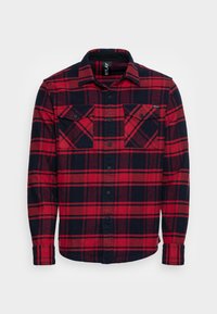 Camicia in flanella a quadri rossa e blu navy con un'apertura frontale a bottoni, due tasche sul petto e colletto. Il tessuto sembra morbido e strutturato.