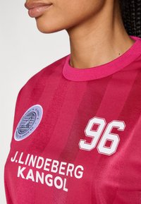 Fuksijas džersija ar teksturētām svītrām, apaļa kakla izgriezums, ar baltu futbola numuru "96" un logotipiem J. Lindeberg un Kangol.