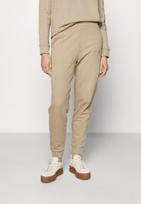Beige Jogginghose aus weichem Stoff, mit elastischem Bund und Bündchen an den Knöcheln, kombiniert mit weißen Sneakers und einem passenden Oberteil.