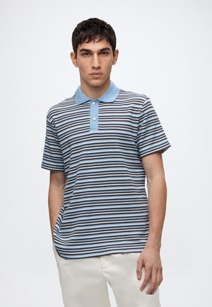 Junger Mann trägt ein blau-braun-weiß gestreiftes Poloshirt mit beiger Hose, steht vor einem schlichten weißen Hintergrund.