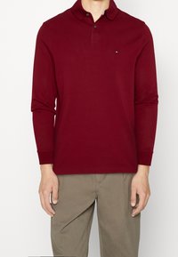 Červená polokošile s dlouhým rukávem a límcem, se třemi knoflíky, malým logem na levé hrudi a žebrovanými manžetami. Kombinováno s béžovými kalhotami.