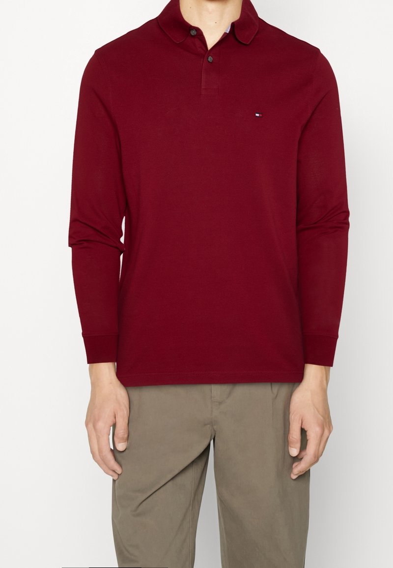 Červená polokošile s dlouhým rukávem a límcem, se třemi knoflíky, malým logem na levé hrudi a žebrovanými manžetami. Kombinováno s béžovými kalhotami.