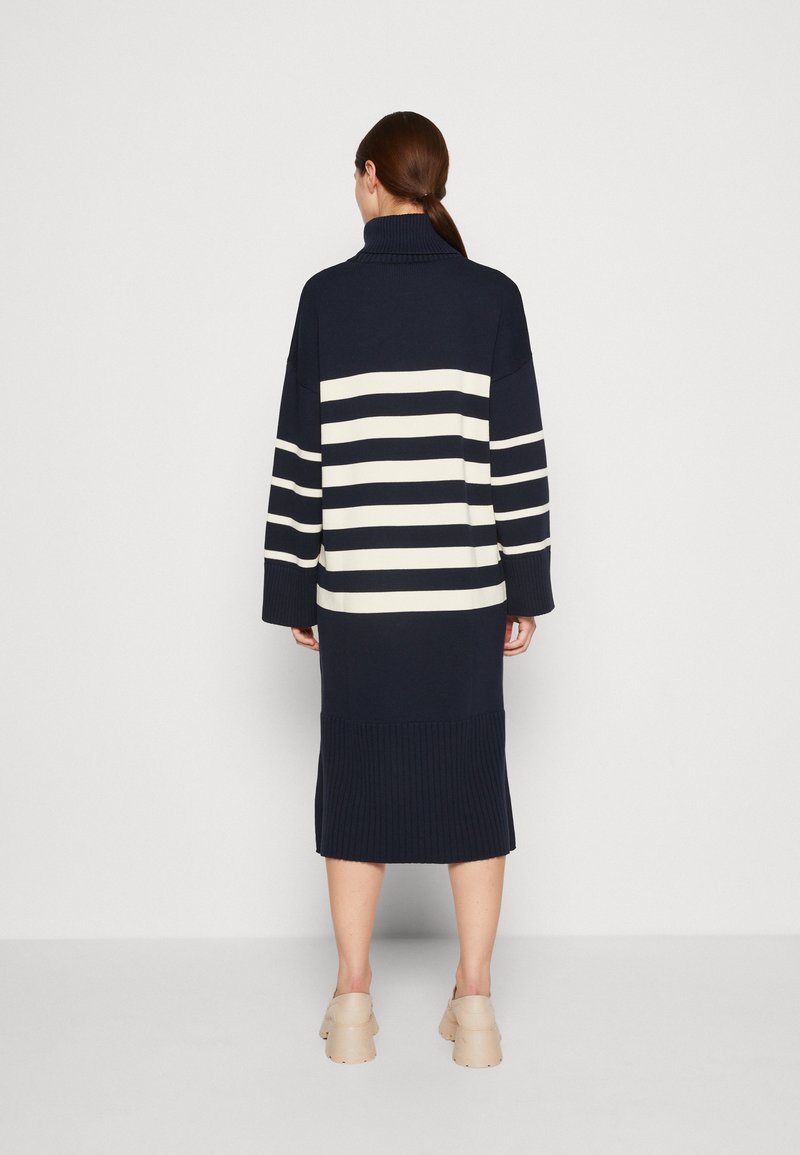 Długa granatowa sukienka swetrowa w białe poziome paski, z prążkowanym dołem i wysokim kołnierzem. Miękka dzianinowa tekstura. Długość poniżej kolan z luźnym krojem.