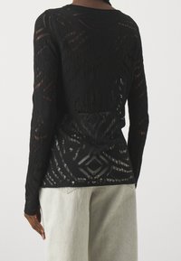 Czarny top z długim rękawem z koronką w geometryczne wzory noszony przez osobę w jasnoszarych spodniach, ukazany z tyłu i od pasa w górę.