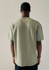 Ljus olivgrön bomulls t-shirt, korta ärmar, avslappnad passform, synlig sömnad på oket, avslutat fåll, som bärs av en person bakifrån.