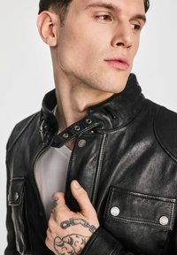 Czarna skórzana kurtka z zapięciem na zatrzaski, srebrnymi detalami i dwiema kieszeniami na piersi. Model ma widoczne tatuaże na dłoni i białą koszulkę pod spodem.