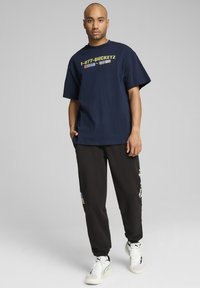 Navy zils īsām piedurknēm t-krekls ar priekšējām grafikām un melnā krāsā adidas biksēm ar uzšuvēm. Apavi ir baltā krāsā ar melniem akcentiem.