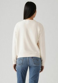 Felpa color crema con scollo rotondo e polsini a costine, abbinata a jeans in denim blu con tasche posteriori dotate di un motivo di cucitura distintivo.