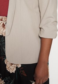 Blazer beige chiaro con una texture liscia, caratterizzato da maniche arrotolate e un semplice revers. Abbinato a una gonna nera con motivo floreale.