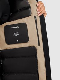 Indersiden af en dunjakke med en blanding af beige og sorte materialer. Indeholder en mærkat, en lynlåslomme og et quiltet mønster.