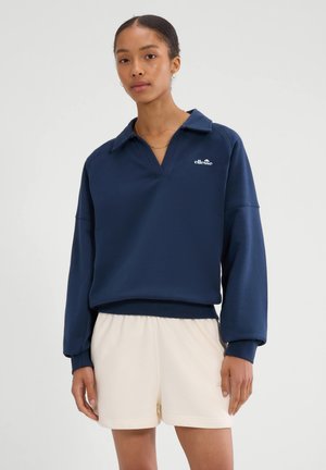 Frau trägt einen marineblauen ellesse-Pullover mit Kragen und cremefarbene Shorts, steht vor einem schlichten hellen Hintergrund.