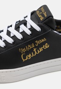 Baskets en cuir noir avec lacets blancs, lettrage doré "Versace Jeans Couture", semelle en caoutchouc, texture lisse, design classique à tige basse.
