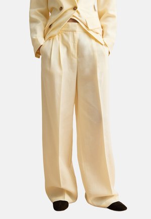 Pantalones de corte suelto color crema con pinzas delanteras, combinados con una chaqueta a juego y zapatos negros de punta.