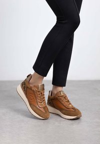 Zapatillas de cuero y ante marrón con acentos de estampado de leopardo, cuello acolchado y suela de goma ligera en contraste. Pantalones negros ajustados visibles.