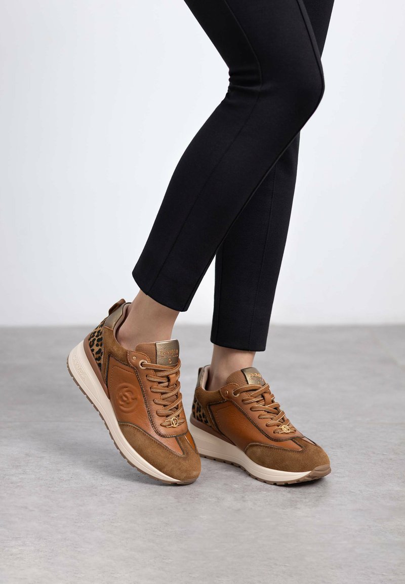 Zapatillas de cuero y ante marrón con acentos de estampado de leopardo, cuello acolchado y suela de goma ligera en contraste. Pantalones negros ajustados visibles.